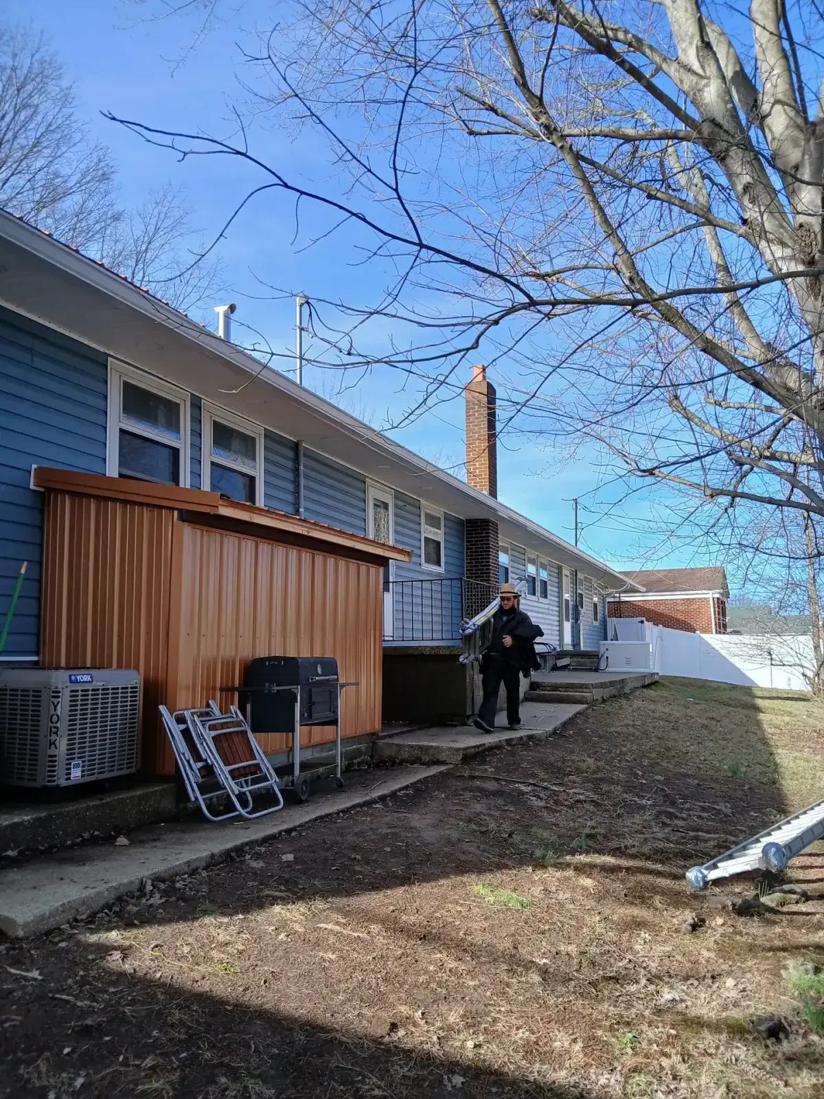 Roofer carrying a ladder past a ranch house with a newly re-roofed rear addition
